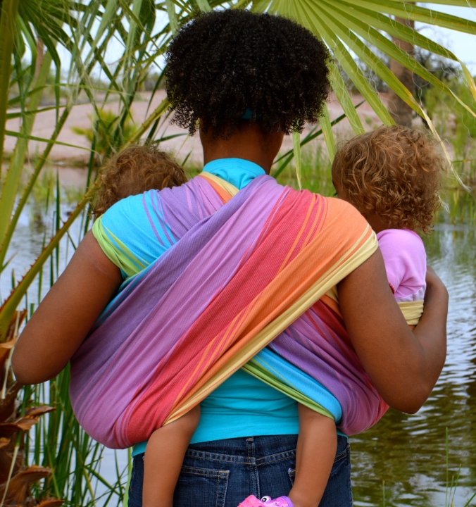 The back view of the tandem hip carry showing the rainbow wrap crossing in an X pattern across the brown skin woman's back. Babies are worn in the wrap one on each hip