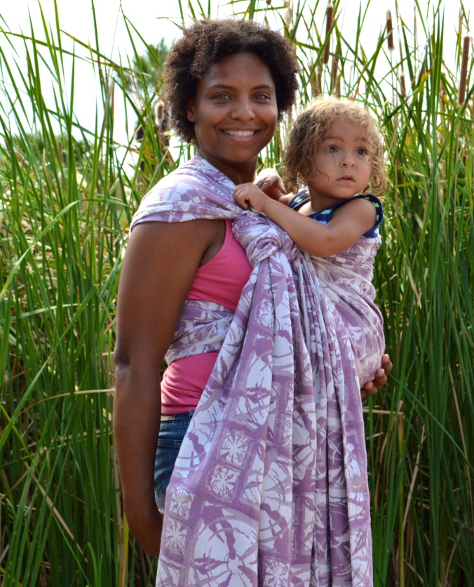 Medium brown natural curly haired momma smiles at the camera while wearing her light brown, blong haired toddler in a front carry in a pale purple and white wrap finished with a slipknot at the shoulder
