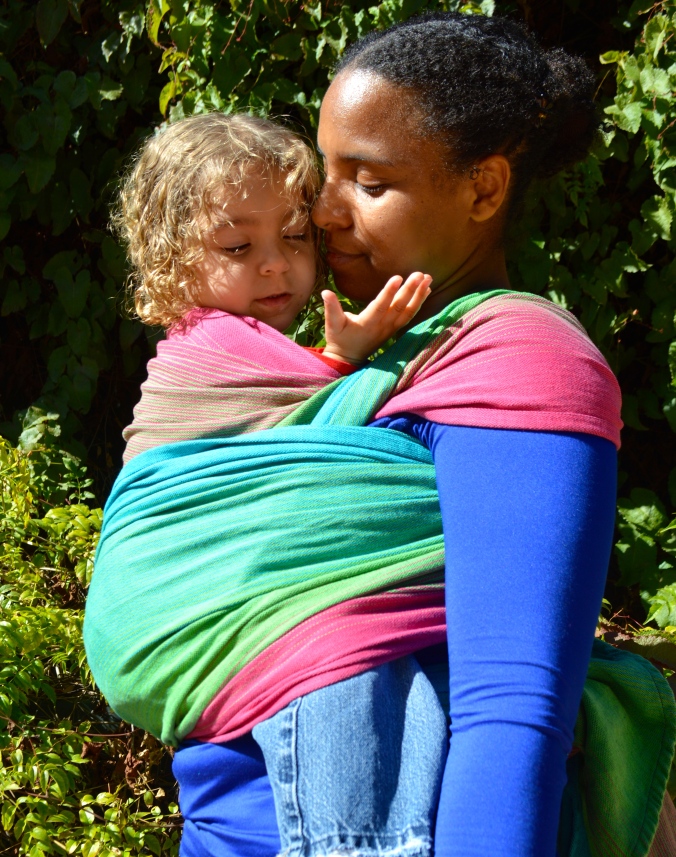 Momma gives kisses to toddler worn in a front carry in magenta, green, pink, and turquoise wrap.