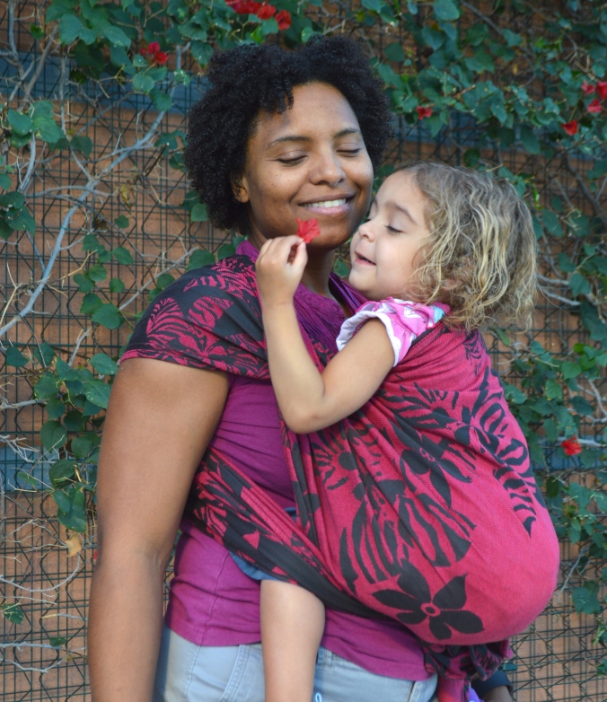 Momma smiles at baby holding magenta flowers in a front carry in a magenta wrap with black flowers