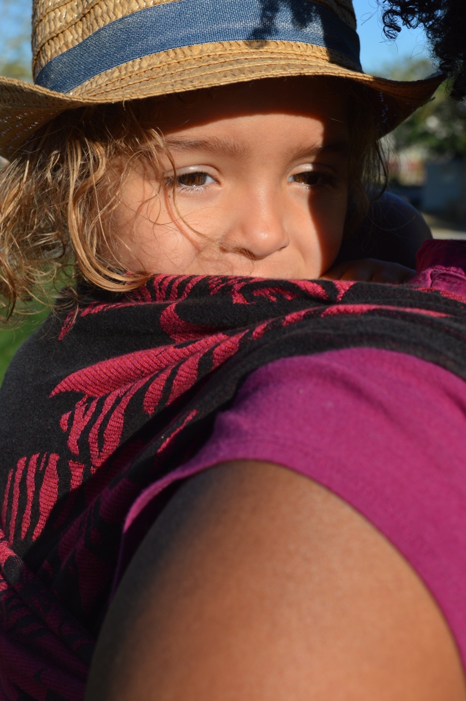 Close up shot of baby's face looking out toward the camera wearing a straw fedora hat. Baby is on Momma's back in a black wrap with magenta flowers