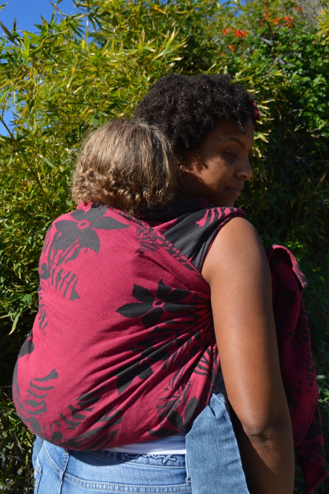 Momma looks back over her shoulder at baby on her back in a magenta wrap with black flowers