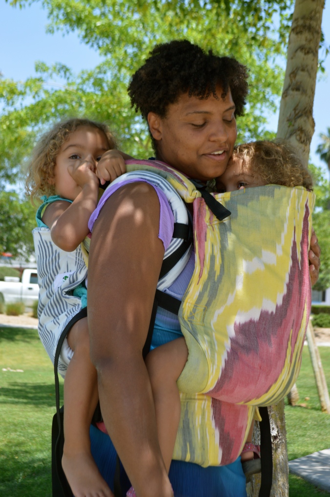 Image shows a medium brown momma wearing a light brown, blond haired toddler on her back in a gray and white diamond print soft structured carrier. The baby worn on her front is hiding, arms tucked inside of the pink, yellow, and white soft structured carrier