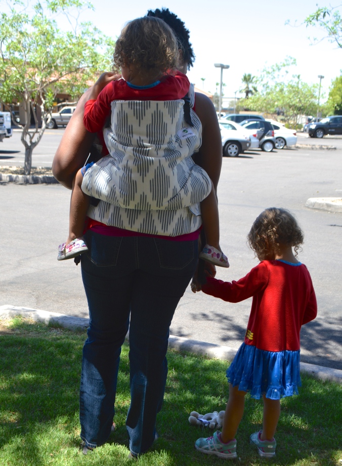 Image shows a back view of a toddler worn in a gray and white diamond print toddler sized soft structured carrier. The woman holds another toddler's hand as they near the street