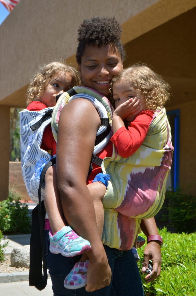 Image shows medium brown woman smiling into the distance while wearing two light brown toddlers. One baby is on her back in a gray and white soft structured carrier. Front baby is worn in a yellow Ikat design with large pink peaks