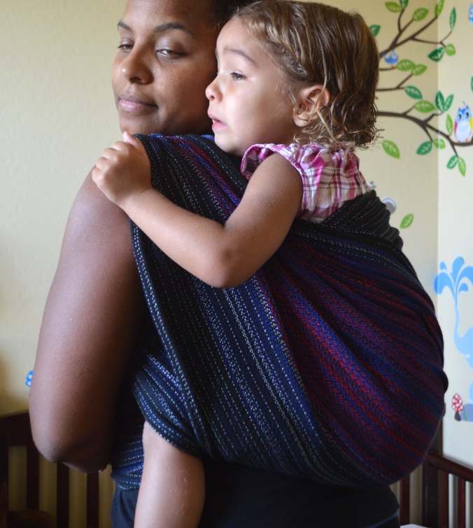 Image of a medium brown woman looking over her shoulder wearing a light brown, blond haired toddler on her back in a black, red, and blue wrap