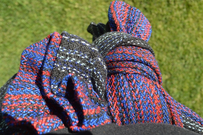 Closeup detail of the weave pattern and colors in the hand woven wrap. The colors transition from black with silver gray stripes to rich red and vibrant blue. The color transitions back to black and gray on the end of the rails. The image is from the point of view of the wearer, looking down at a large knot