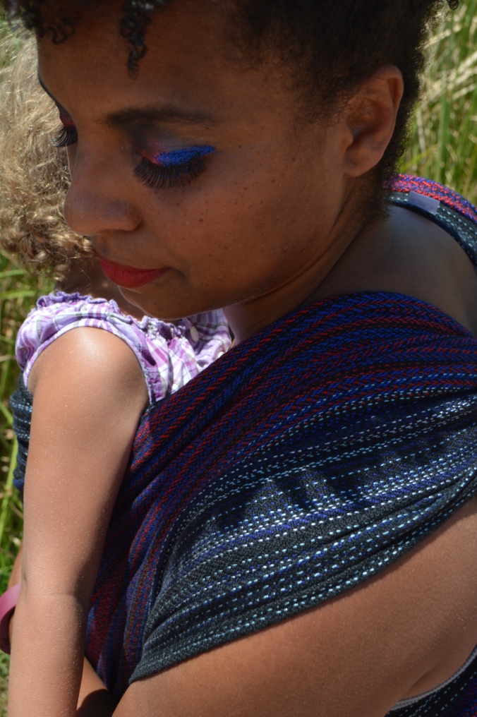 Close up view, over the shoulder of the wearer showing the black, red, and blue of the wrap on her shoulder and the baby's arm in view. Her eye shadow is red and blue glitter with long black eyelashes.