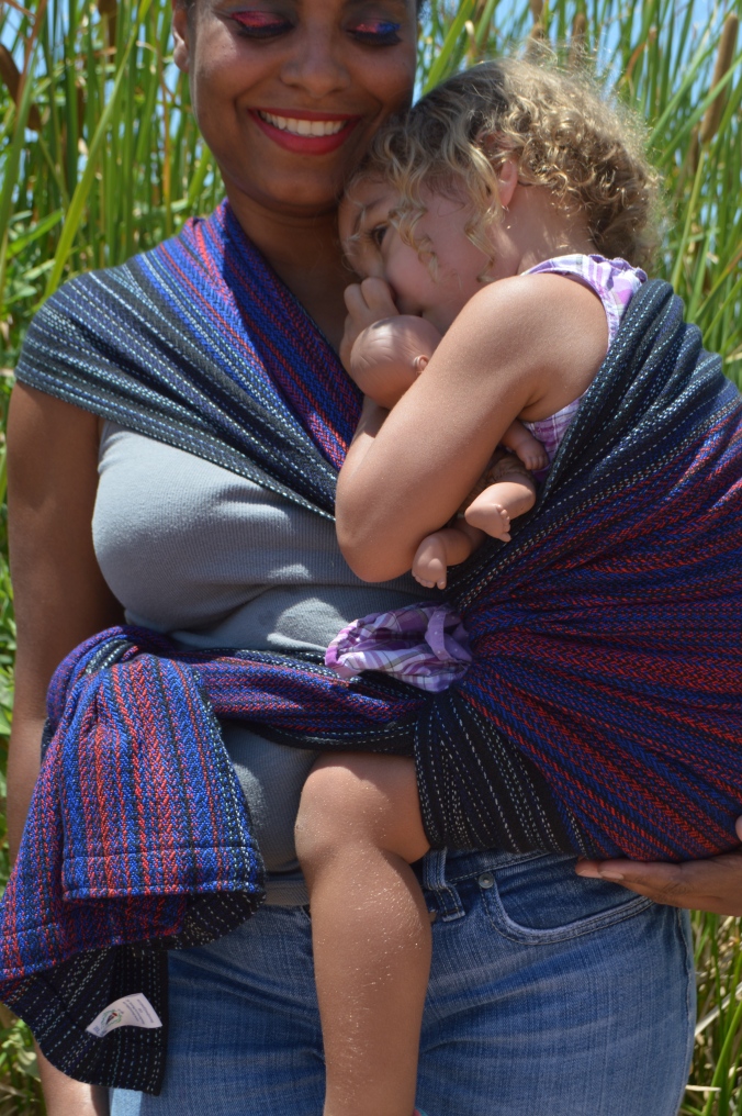 Image shows a medium brown african american momma cuddled close with light brown multiracial blond haired toddler in a Hip carry. They are standing in front of tall green reeds wearing a black red, and vibrant blue hand woven wrap. Baby is snuggled in close, thumb in mouth, heavy lidded ready for nap