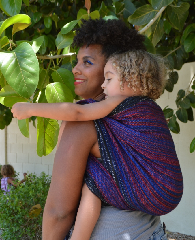 Image of a medium brown woman looking over her shoulder wearing a light brown, blond haired toddler on her back in a black, red, and blue wrap. Momma and baby are looking at leaf patterns in the trees above