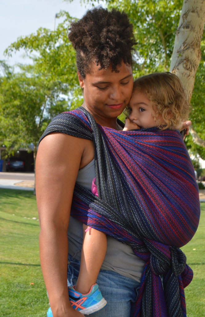 Image shows a medium brown african american momma cuddled close with light brown multiracial blond haired toddler in a front carry. They are standing in front of tall green reeds wearing a black red, and vibrant blue hand woven wrap. Baby is snuggled in close with thumb in her mouth