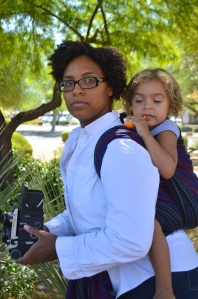Image of a medium brown woman looking over her shoulder wearing a light brown, blond haired toddler on her back in a black, red, and blue wrap. The woman wears glasses and is dressed as Peter Parker in a white shirt holding a vintage Mamyia medium format camera