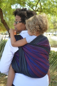 Image of a medium brown woman looking over her shoulder wearing a light brown, blond haired toddler on her back in a black, red, and blue wrap. The woman wears glasses and is dressed as Peter Parker in a white shirt
