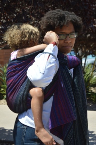 Image of a medium brown woman looking over her shoulder wearing a light brown, blond haired toddler on her back in a black, red, and blue wrap. The woman wears glasses and is dressed as Peter Parker in a white shirt. The woman is reaching for her glasses as if to take them off