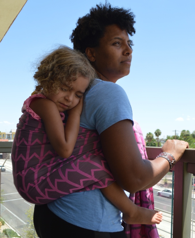 Image of a medium brown woman with natural curly hair staring off in the distance wearing a light brown toddler in a gray woven wrap with pink zig-zag pattern woven wrap