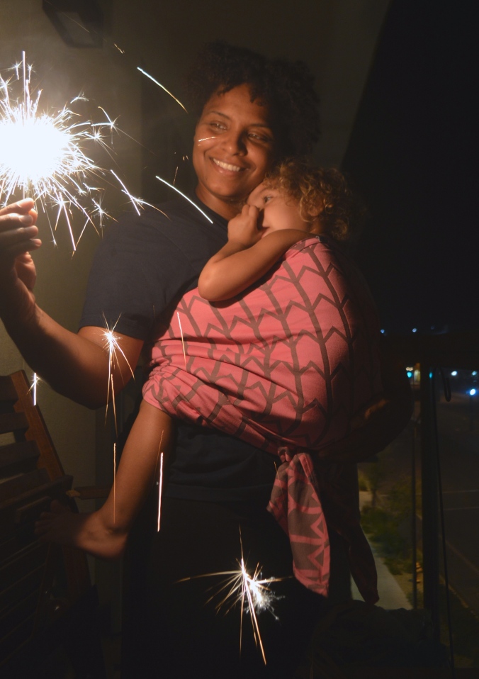 Image of a woman wearing a scared toddler in a front carry as they both look at the sparkler fizzling bright at night