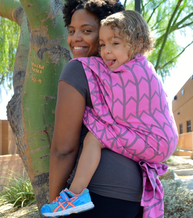 Image os a medium brown woman with natural hair wearing a light brown toddler with curly blonde air on her back in a vibrant pink woven wrap with gray zig-zag pattern