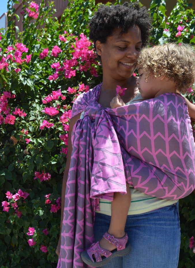 Image of a medium brown woman with natural curly hair wearing a light brown toddler with blonde hair in a gray woven wrap with pink zig-zag pattern. Momma is looking down at the toddler who is holding a hot pink flower